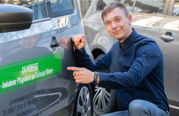 Junger Mann zeigt auf das Dienstauto des Pflegedienstes KAI VIEREGGE